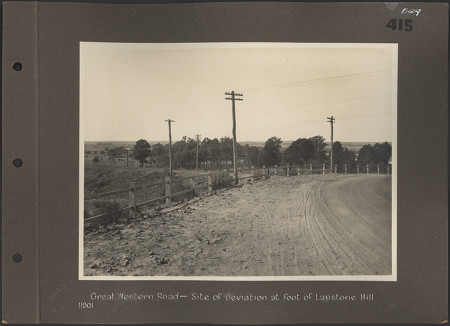 A black and white photograph stuck to a scrapbook page showing a dirt road lined with a fence and telegraph poles. The photo is labelled ‘Great Western Road- site of deviation at foot of Lapstone Hill’. The left side of the page has binder holes.